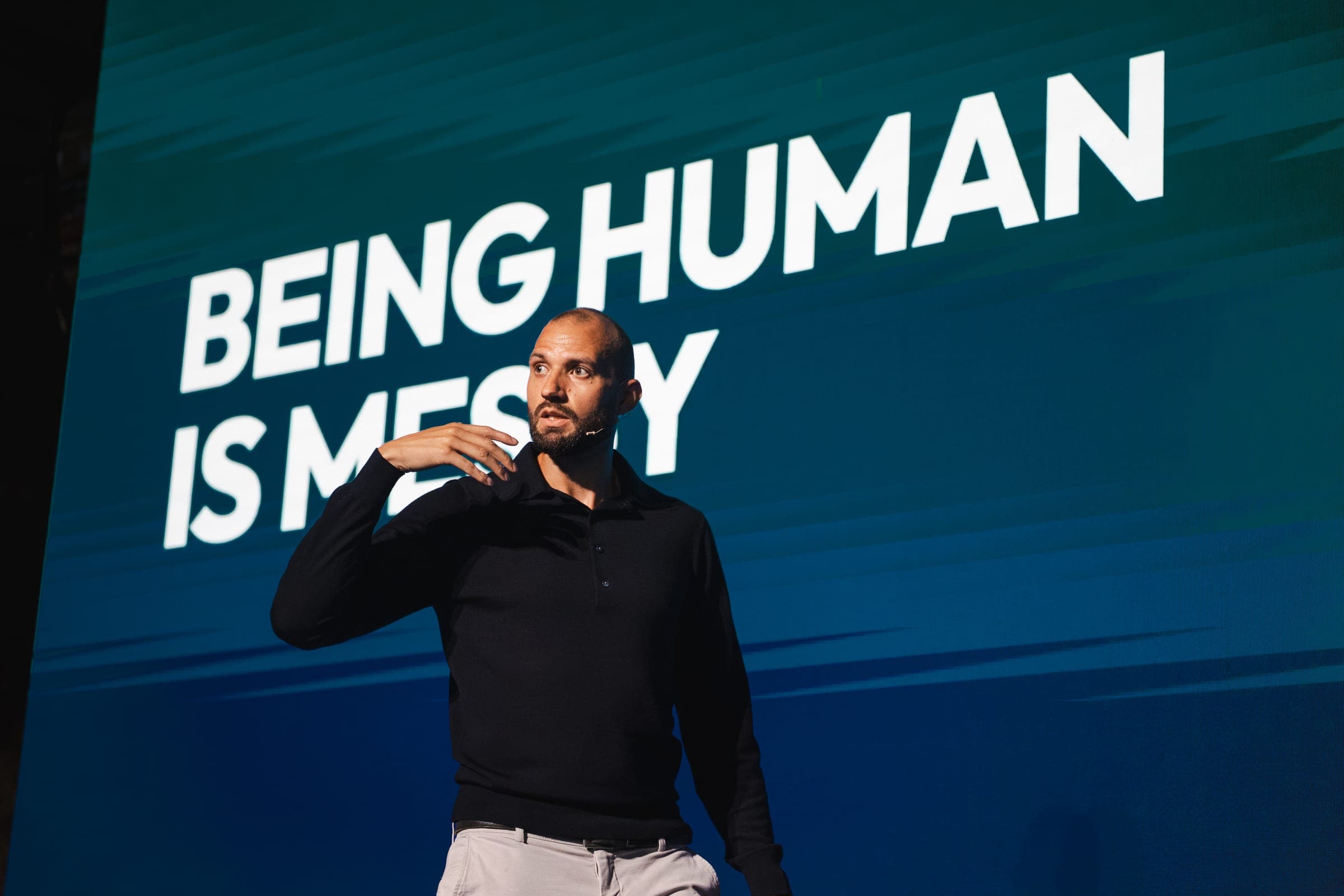 A speaker on stage in front of a slide that reads "Being human is messy"